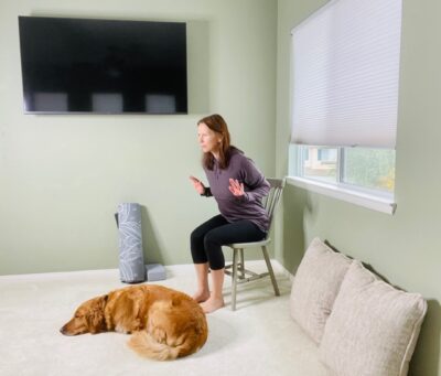 A woman seated in a chair next to a window with her dog near her practicing a chair yoga pose of modified low push up with her elbows bent near her sides and the wrists extended.