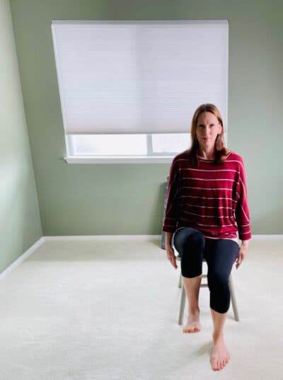 A woman seated in a chair near a window with one knee lifted in the air for chair yoga low lunge pose.