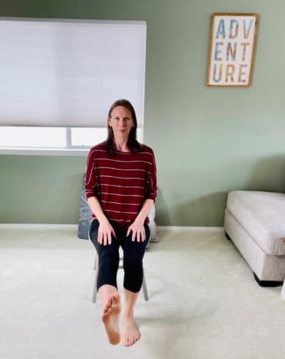 A woman seated in a chair near a window for chair yoga with one knee extended.