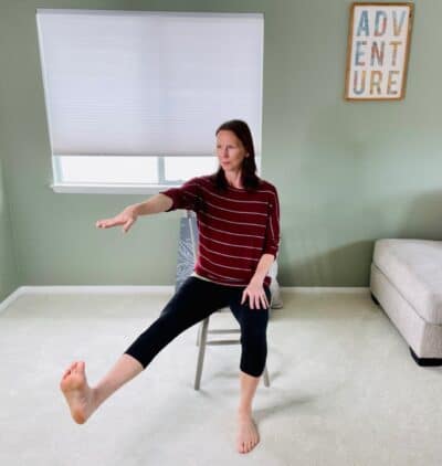 A woman seated in a chair near a window with one knee extended and out to the side and reaching one hand forward for chair pose hand to big toe variation.