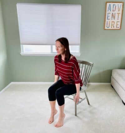 A woman seated in a chair near a window practicing modified chair pose with heels raised and arms by her sides.