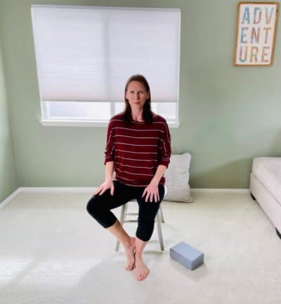 A woman seated in a chair near a window practicing the chair yoga tree pose with her toes on the ground and her heel resting on the inside of the opposite shin.