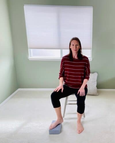 A woman seated near a window practicing a modified tree pose seated using a yoga block.