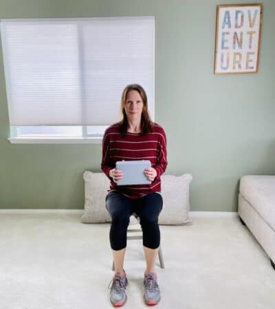 A woman seated in a chair near a window holding a yoga block with both hands in front of the body.