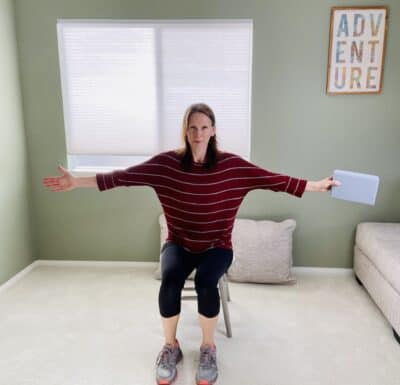 A woman seated in a chair near a window with both arms stretched out wide with one hand holding a yoga block.