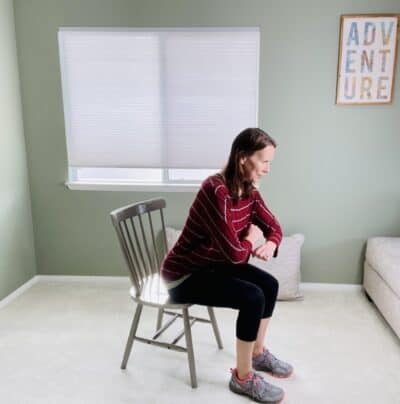 A woman seated in a chair near a window doing forearm circles with a hip hinge exercise.