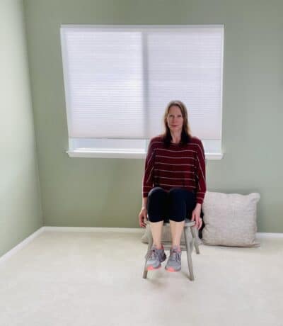 A woman seated in a chair near a window with her hands slightly braced on the chair and both knees raised up toward the sky.