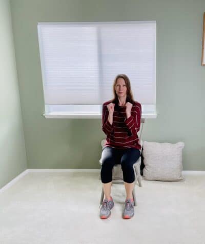 A woman seated in a chair near a window with both elbows bent and hands in a loose fist preparing for seated cross punch exercise.