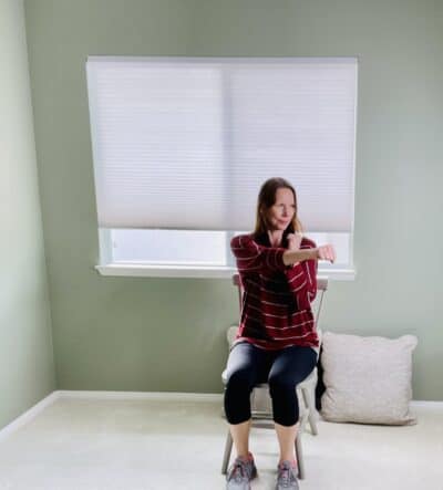 A woman seated in a chair near a window with one arm near the body and the other punching across the body for seated cross punch exercise.