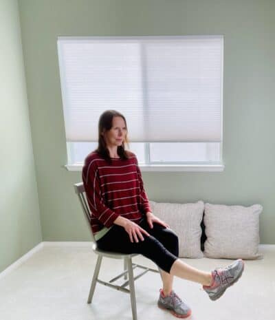 A woman seated in a chair near a window with one knee extended and performing ankle range of motion exercises.