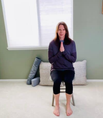 A woman seated in a chair near a window with palms together and hands at her chest for chair yoga pose, hands to heart pose.