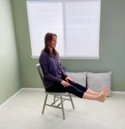 A woman seated in a chair near a window to practice chair yoga pose, staff pose, with both knees extended and toes pointed toward the sky.