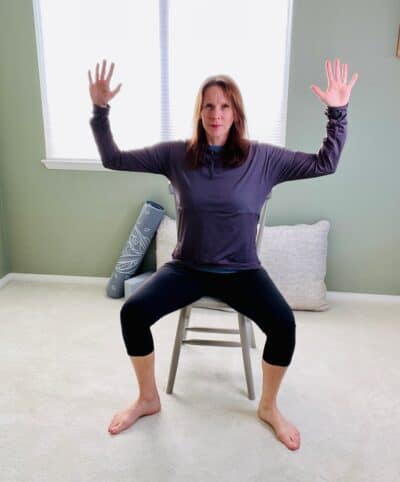 A woman seated in a chair near a window practicing chair yoga pose, goddess pose with her arms in cactus arms and both hips externally rotated and planted on the floor.