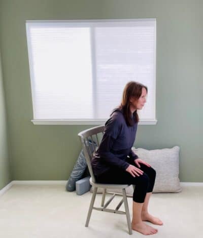 A woman seated in a chair next to a window practicing chair yoga pose, the hip hinge.