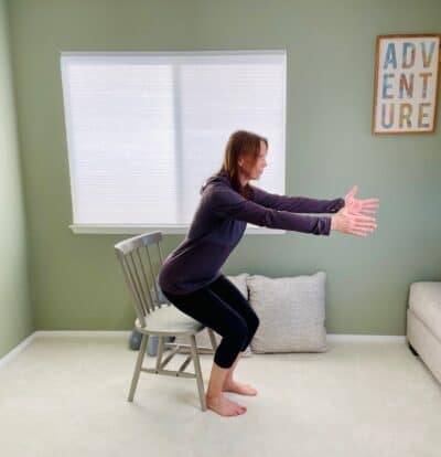 A woman in a room near a window practicing chair yoga chair pose with her arms extended in front of her and her hips lifted off the seat of the chair.