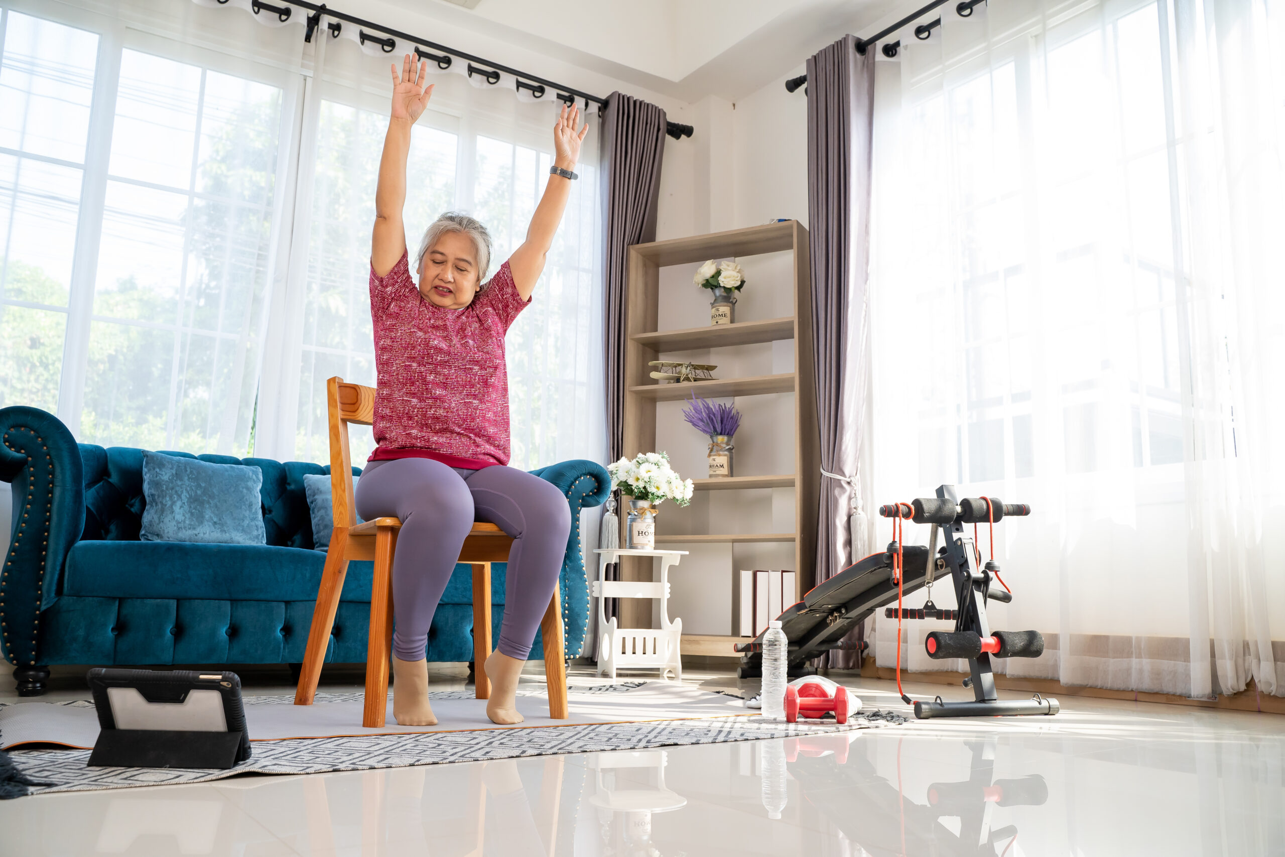 An older woman seated in a chair in her living room surrounded by some exercise equipment with her arms raised up in the air for chair exercises.