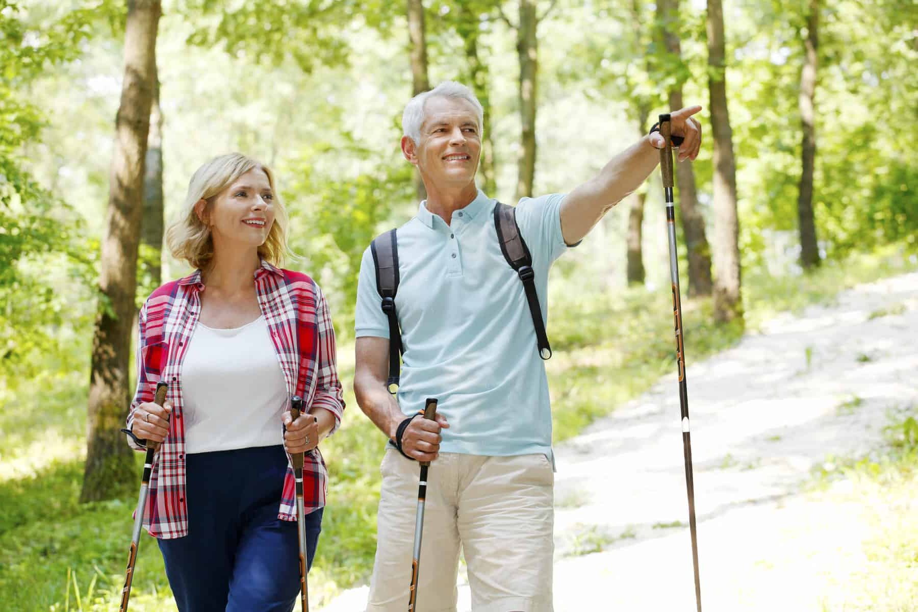 An older couple with walking poles taking a hike on a sunny tree lined trail.