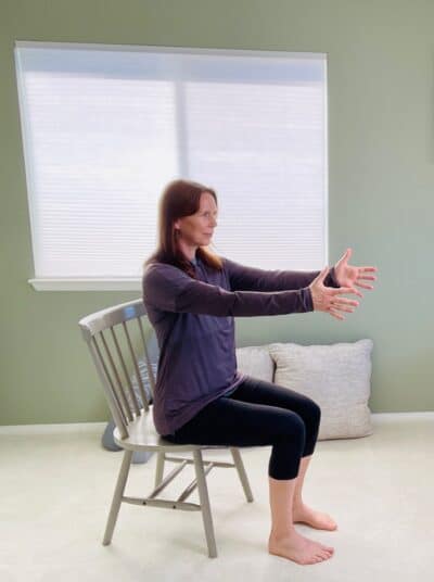 A woman seated in a chair near a window with both arms stretched out in front of the body in preparation for a seated row exercise.