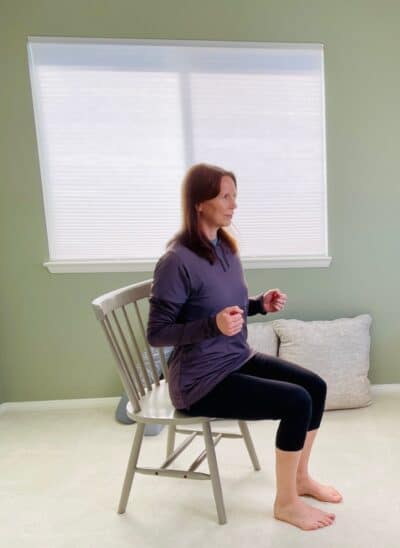 A woman seated in a chair near a window with both elbows bent and arms pulled towards the rib cage at the end of a seated row exercise.