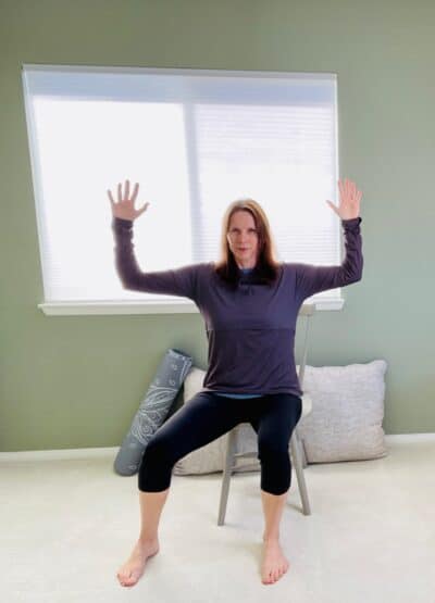 A woman seated in a chair near a window for chair yoga with both arms lifted into Cactus Arms and the right hip externally rotated.