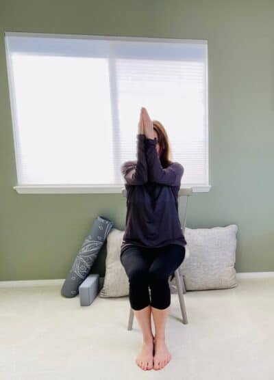 A woman seated in a chair near a window with both arms lifted and elbows and palms together for chair yoga pose.