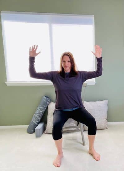 A woman seated in a chair near a window practicing chair yoga pose cactus arms with the left leg in external rotation.