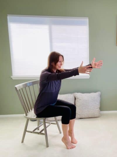 A woman seated in a chair near a window in chair yoga pose, chair pose, with heels elevated and both arms reaching forward.