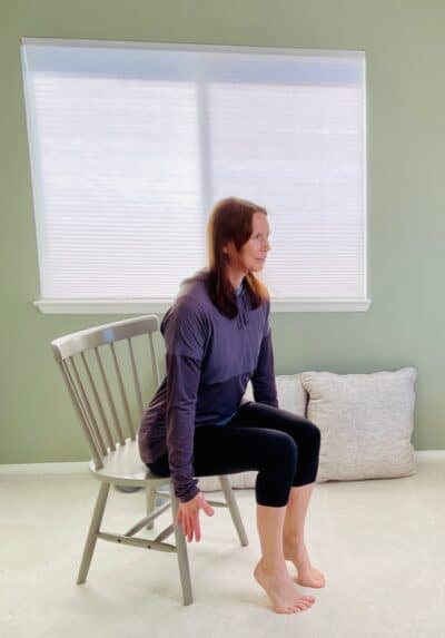 A woman seated in a chair near a window in chair yoga pose, hip hinge with heels raised and arms by her sides.