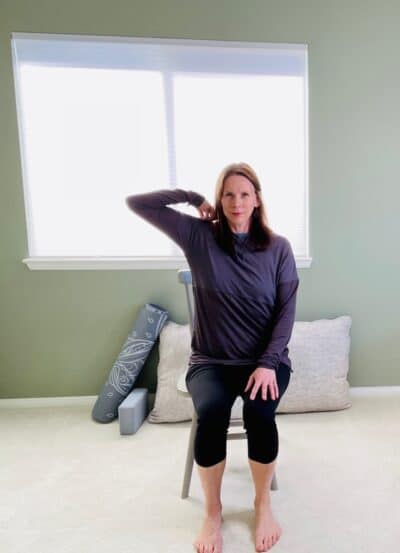 A woman seated in a chair near a window with on elbow bent and the hand resting on the shoulder for lateral side stretch.