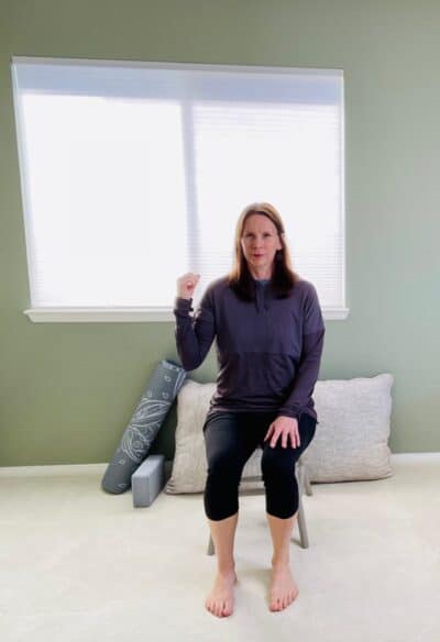 A woman seated in a chair near a window with one elbow bent and the hand facing the sky in preparation for a lateral side stretch.