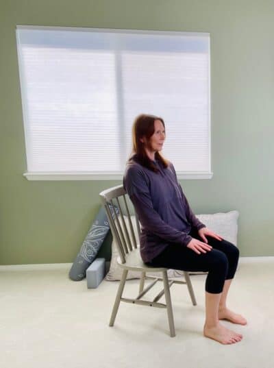 A woman seated in a chair by a window practicing chair yoga pose, boat pose, with both feet planted on the floor and the trunk hinging posteriorly.