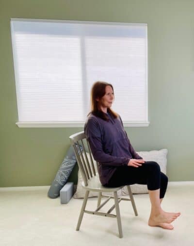 A woman seated in a chair next to a window practicing chair yoga pose, boat pose with one knee lifted.