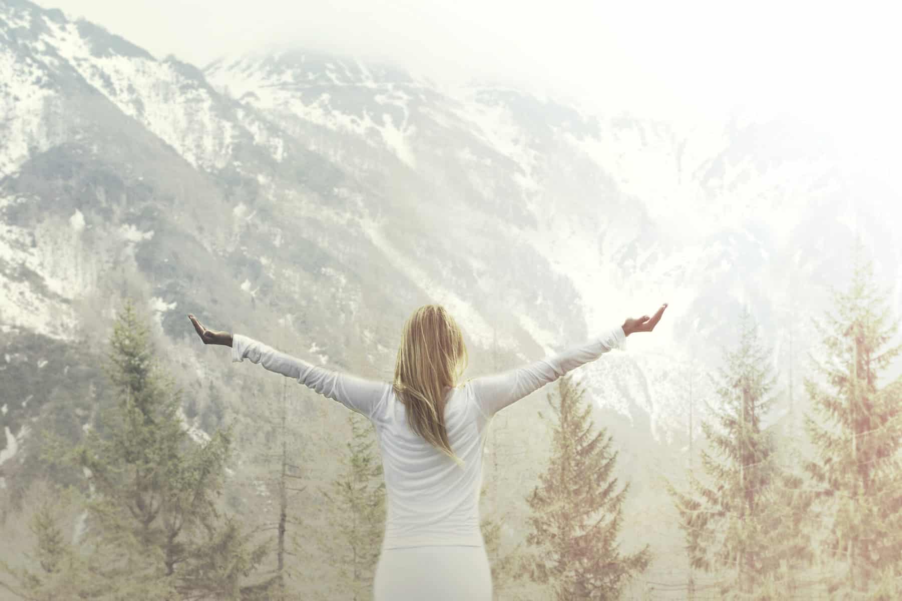 A woman with long hair facing a majestic mountain and taking a deep breath with both of her arms stretched out wide with teh palms facing up.