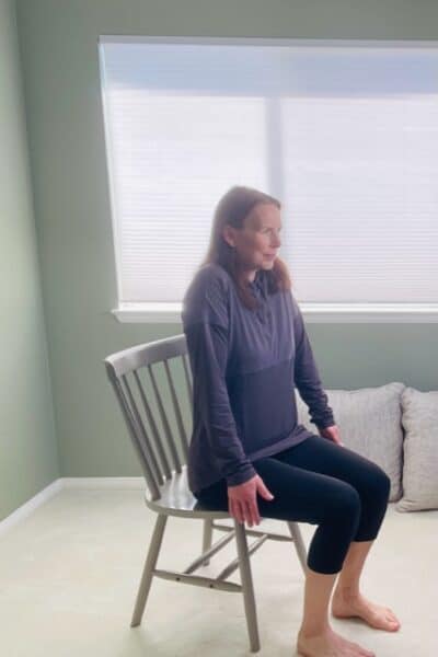 A woman seated in a chair near a window with her shoulders raised up toward her ears for a shoulder shrug exercise.