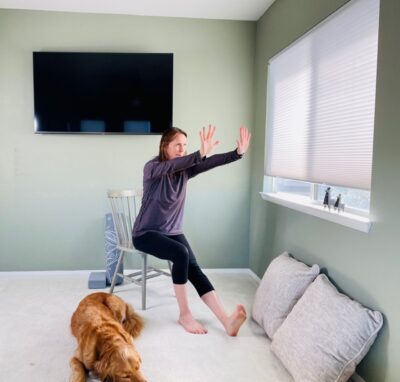 A woman seated in a chair by a window hinged forward from the hips with one knee extended and both arms in front of the body with the palms forward and wrists extended for Modified Downward Facing Dog Chair Yoga Pose.