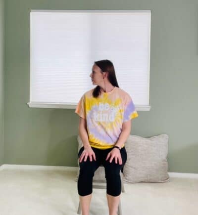 A woman seated in a chair near a window looking over her right shoulder for a seated neck rotation exercise.