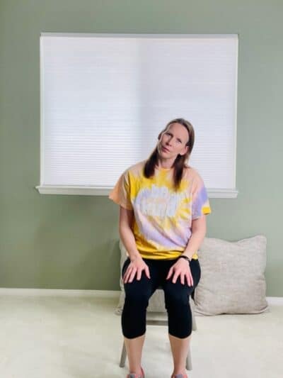 A woman seated in a chair near a window with her left ear towards her left shoulder for a lateral neck stretch.