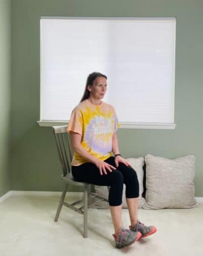 A woman seated in a chair near a window with her toes lifted and her heels on the ground for a seated dorsiflexion exercise.