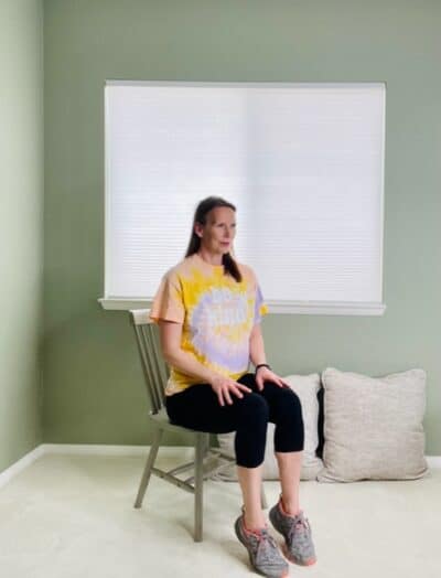 A woman seated in a chair near a window with her toes on the floor and her heels lifted for a seated heel raise.