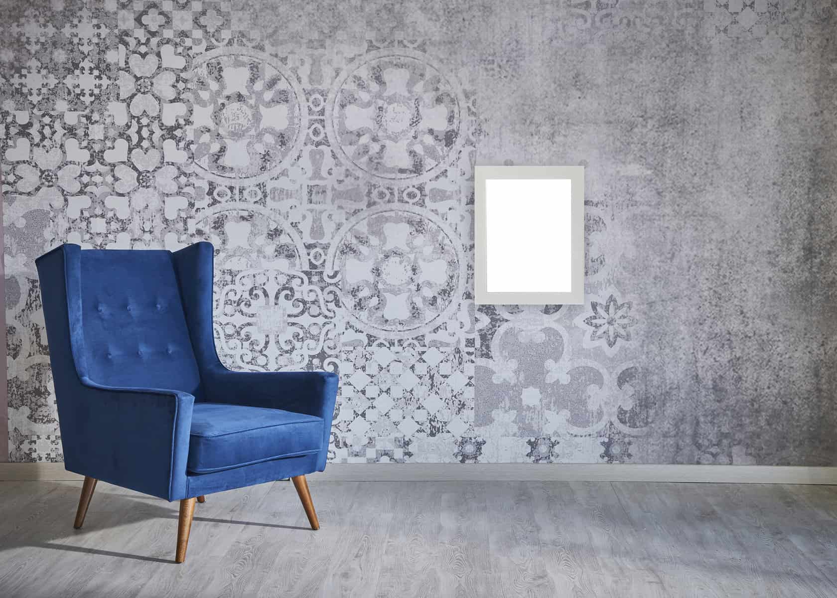 A bright blue armchair on a wood floor in a room with wallpaper covered walls with a rectangle mirror.