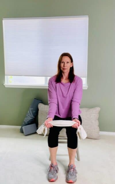 A woman seated in a chair holding a towel with each hand with the elbows extended for a seated chair exercise.