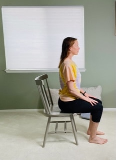 A woman seated in a chair near a window practicing cow pose with her spine elongated and slightly arched.