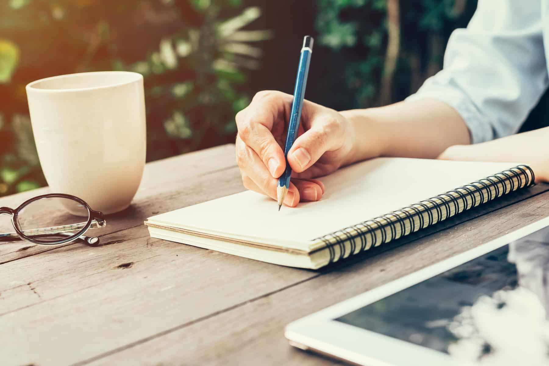 A cropped image of a table with a candle and a pair of glasses on it with a woman's hands holding a pen while she writes in a journal for a mindfulness practice.