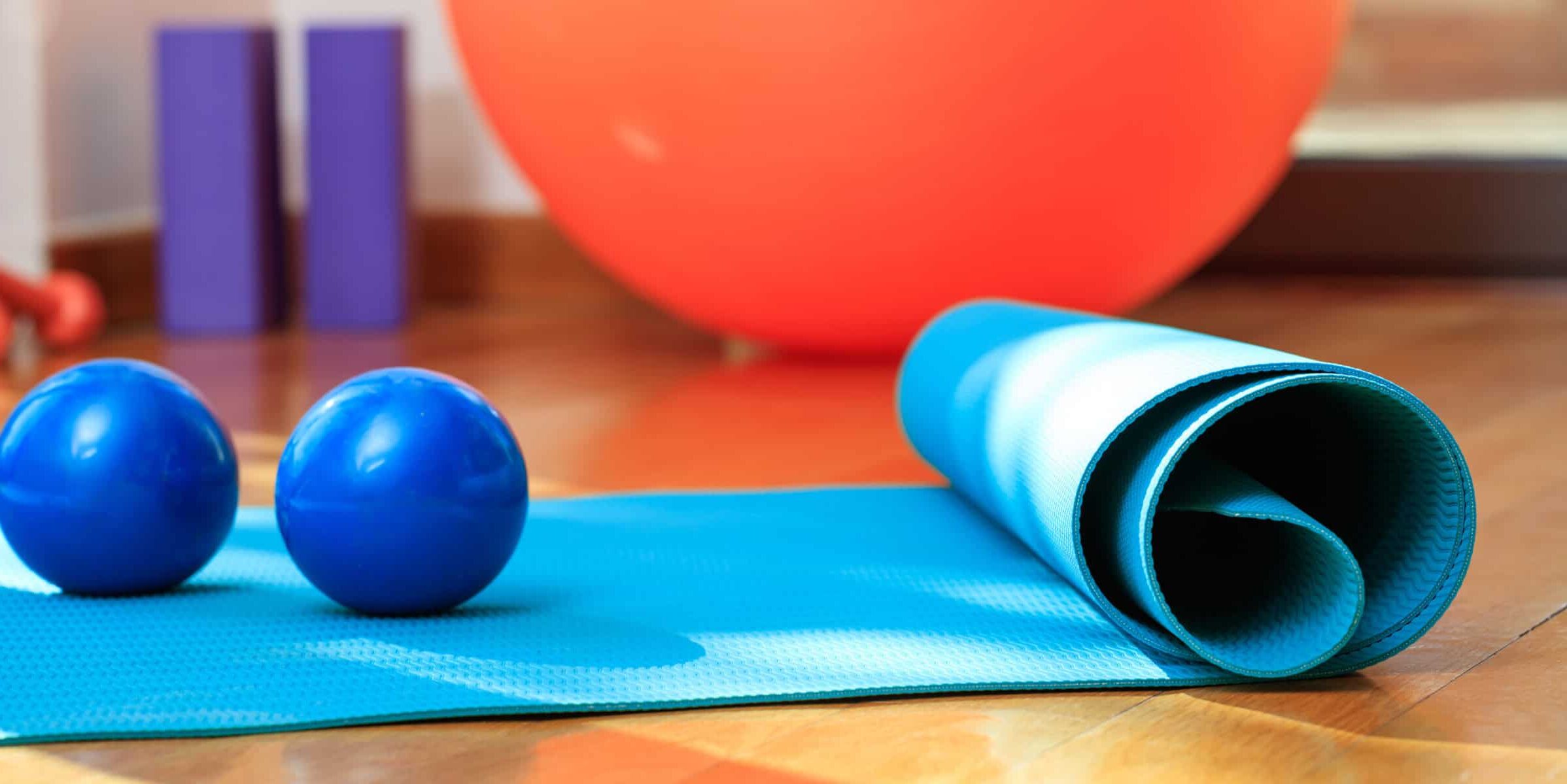 A cropped image of a partially rolled up blue yoga mat on a wood floor with a large orange ball in the background and two smaller blue exercise balls.