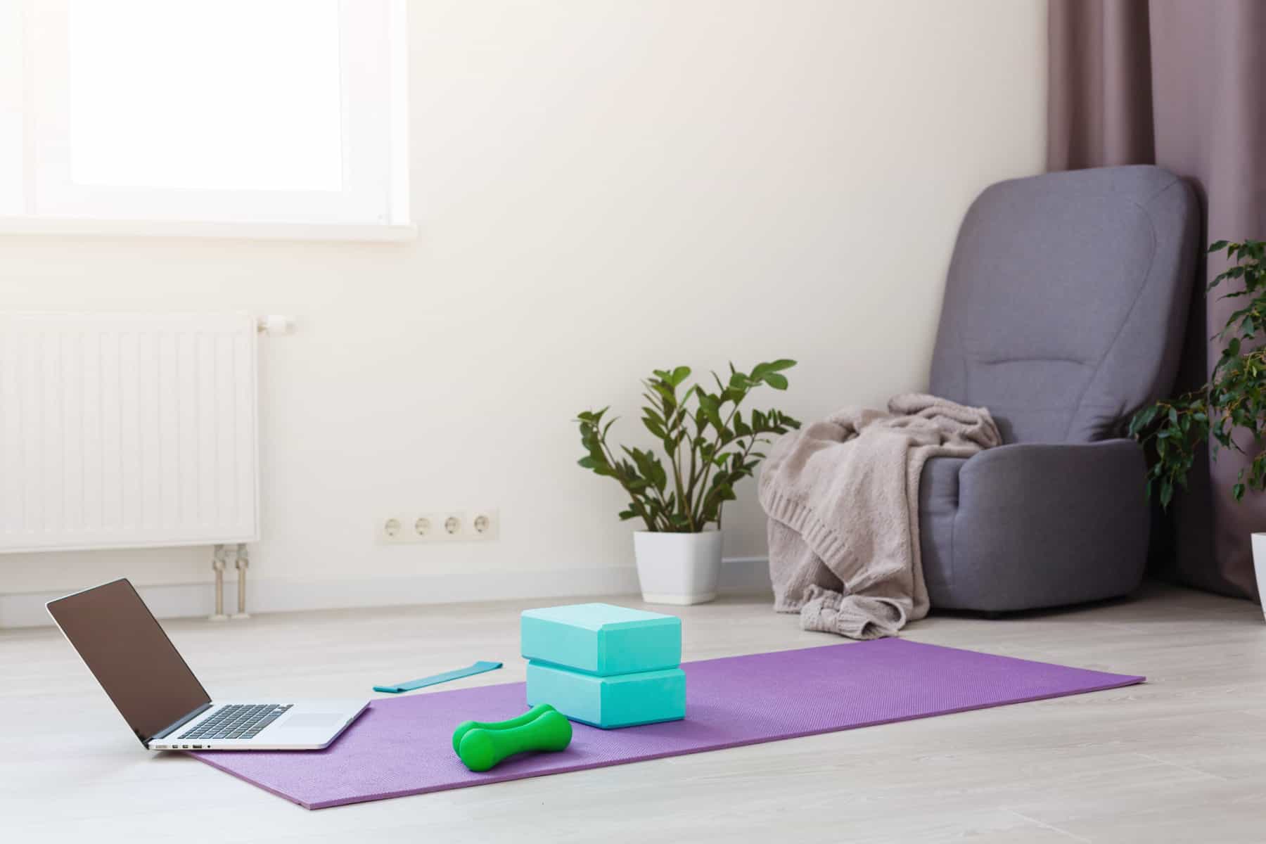 A image of a living room with a gray chair and a purple yoga mat with a lap top, hand weights, yoga blocks, and an exercise band for at home exercise equipment.