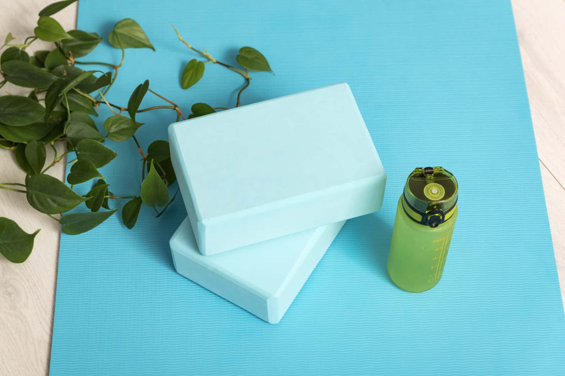 A cropped image of a bright blue yoga mat with two light blue yoga blocks, a green water bottle, and a plant on a light wood floor.