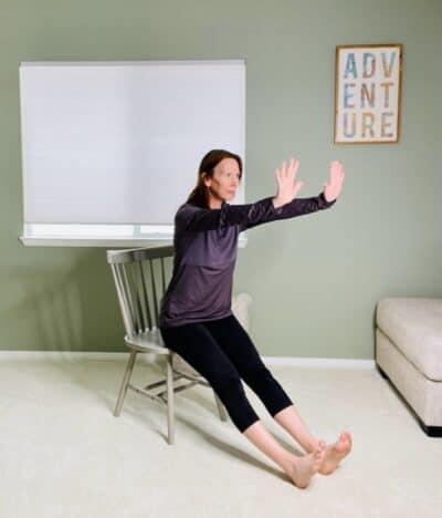 A woman seated in a chair for chair yoga pose Downward Facing dog with both legs extended in front of the body and arms forward with the fingers back towards the face.