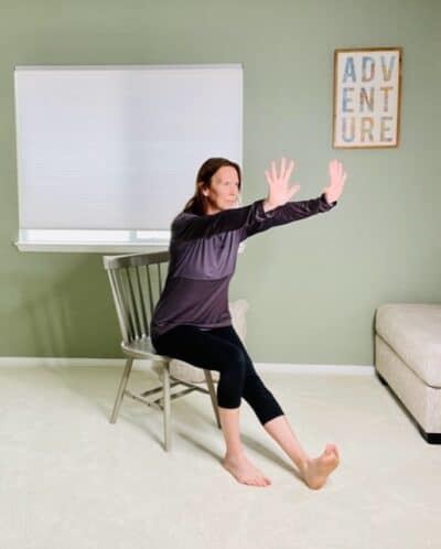 A woman seated in a chair for chair yoga with one leg extended and both arms raised in front of the body with the fingers towards the face for Downward Facing Dog variation.