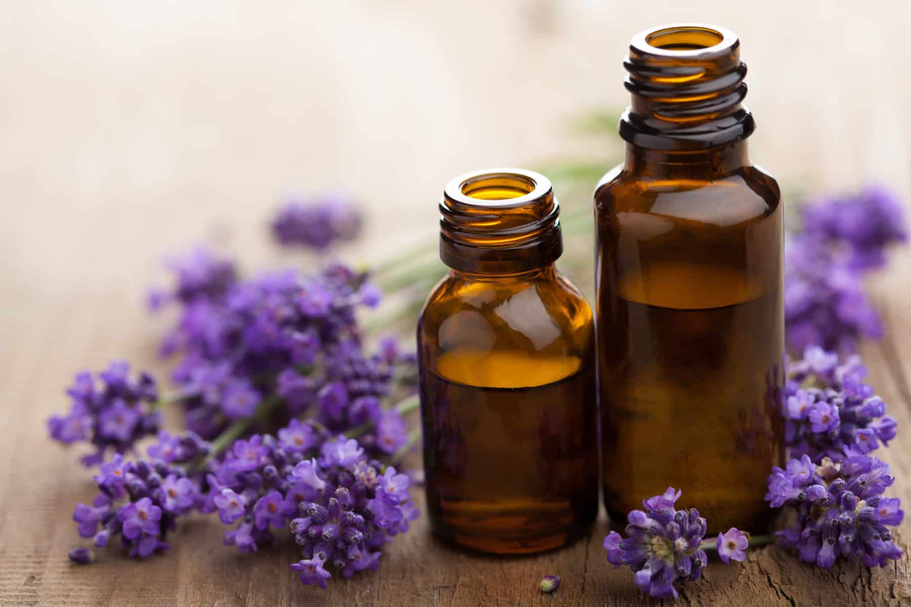 A close up of two bottles of essential oils with purple lavender flowers next to them on a wooden table.