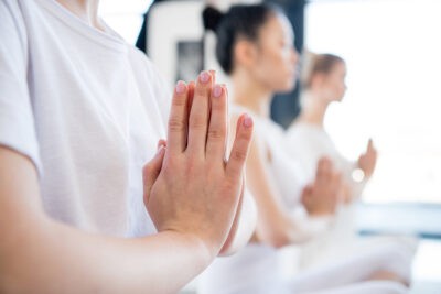 A cropped image of three people in a yoga class with their hands together at their heart for meditation.